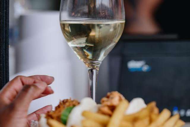 Close-up of a hand holding a white wine glass with fries and food on a plate nearby.
