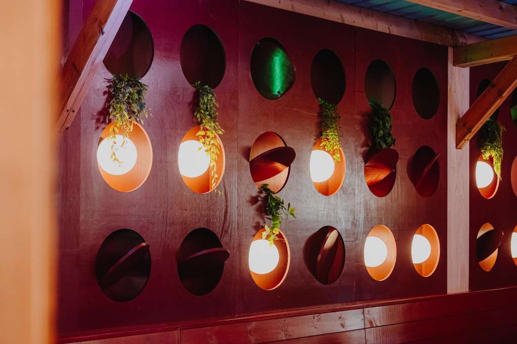 Decorative wall installation with illuminated circular cutouts, trailing greenery, and sculptural red elements against burgundy backdrop.