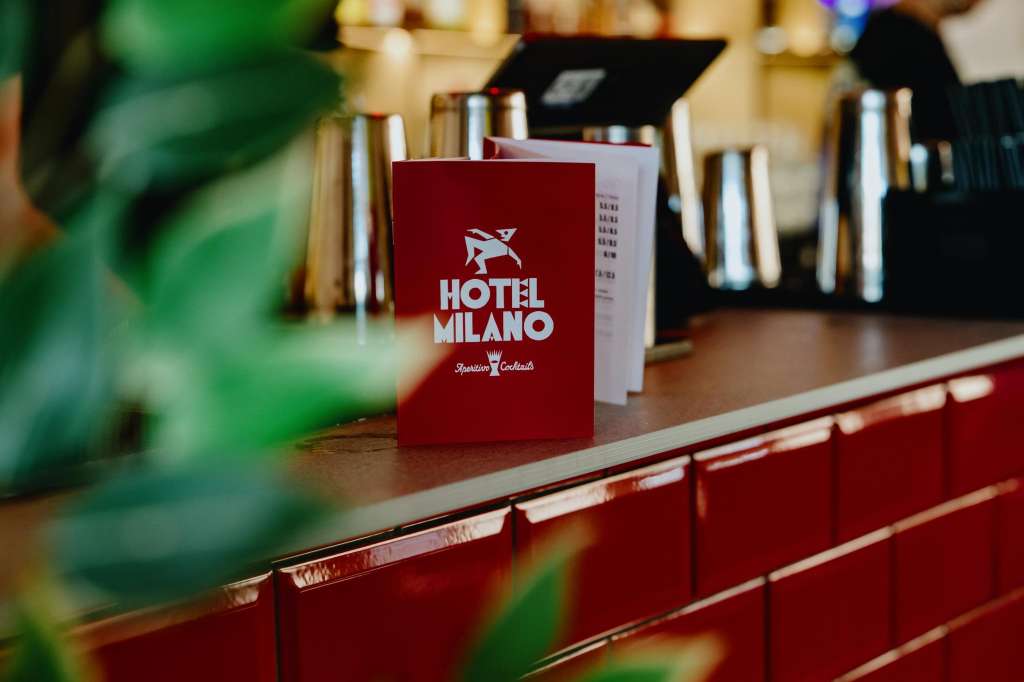 Hotel Milano Bar menu card on red counter with glasses and bar seating in background.