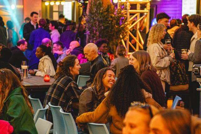 Industrial venue with crowd seated at tables, colorful stage lighting, yellow steel trusses, and performance setup.