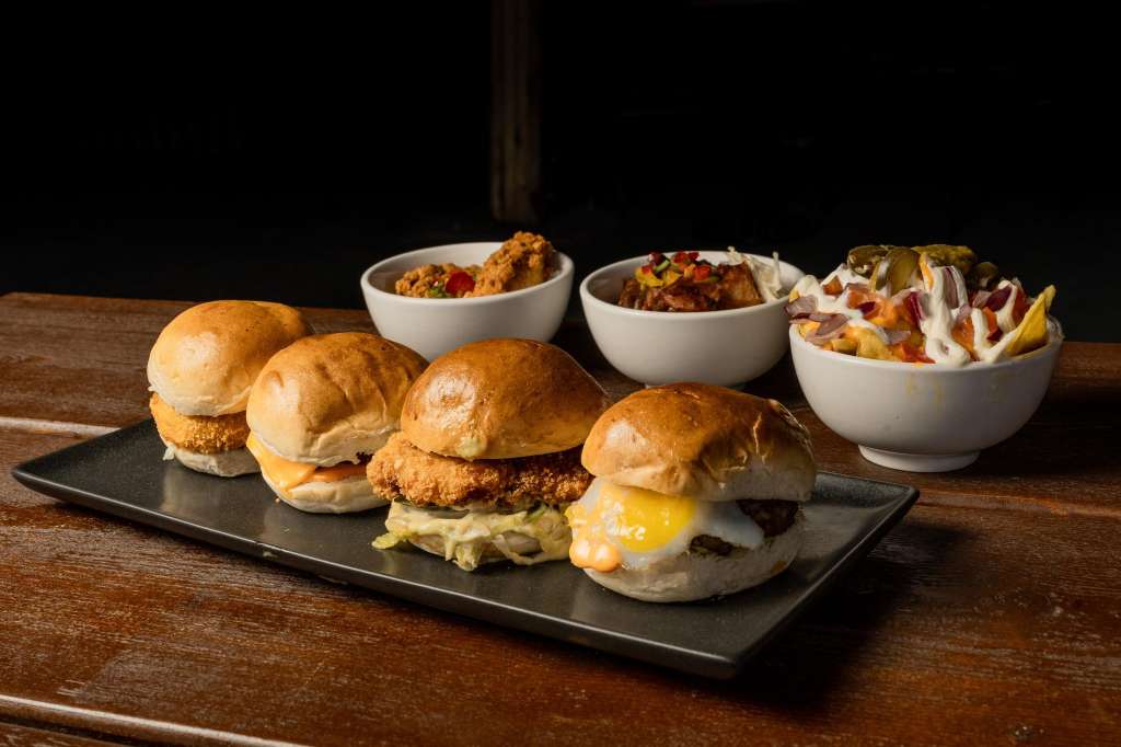 Close-up of fried chicken sliders and side dishes in white bowls on a dark slate plate.