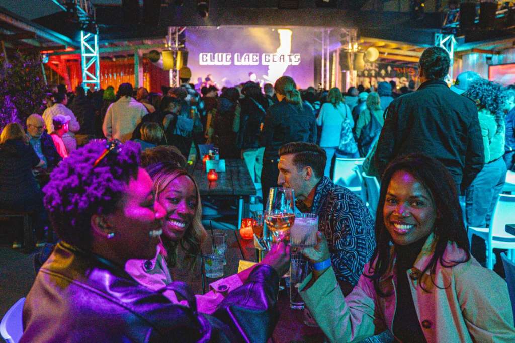 Large indoor venue with crowd enjoying evening event under stage lighting, purple and blue ambient lighting, industrial truss ceiling.