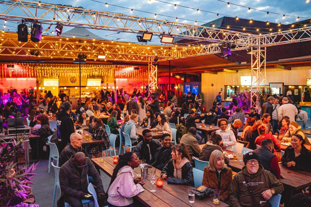 Full venue hire with large crowd seated at tables under industrial truss, colorful stage lighting, and string lights overhead.