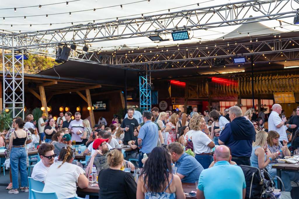 Outdoor venue with industrial truss rigging, string lights, and large crowd mingling at tables under covered structure.