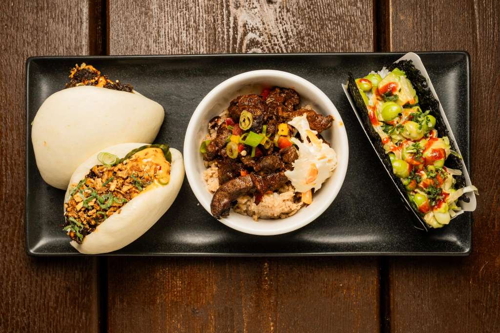 Black rectangular plate with Asian-inspired dishes: steamed buns, braised meat bowl, and vegetable-topped noodles on wooden surface.