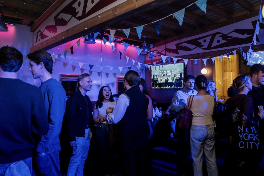 Large indoor space with guests mingling under blue and pink lighting, bunting, and exposed wooden beams.