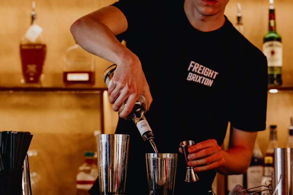 Bartender in black Freight Brixton shirt mixing a cocktail behind a bar with illuminated bottle shelving and red tile front.
