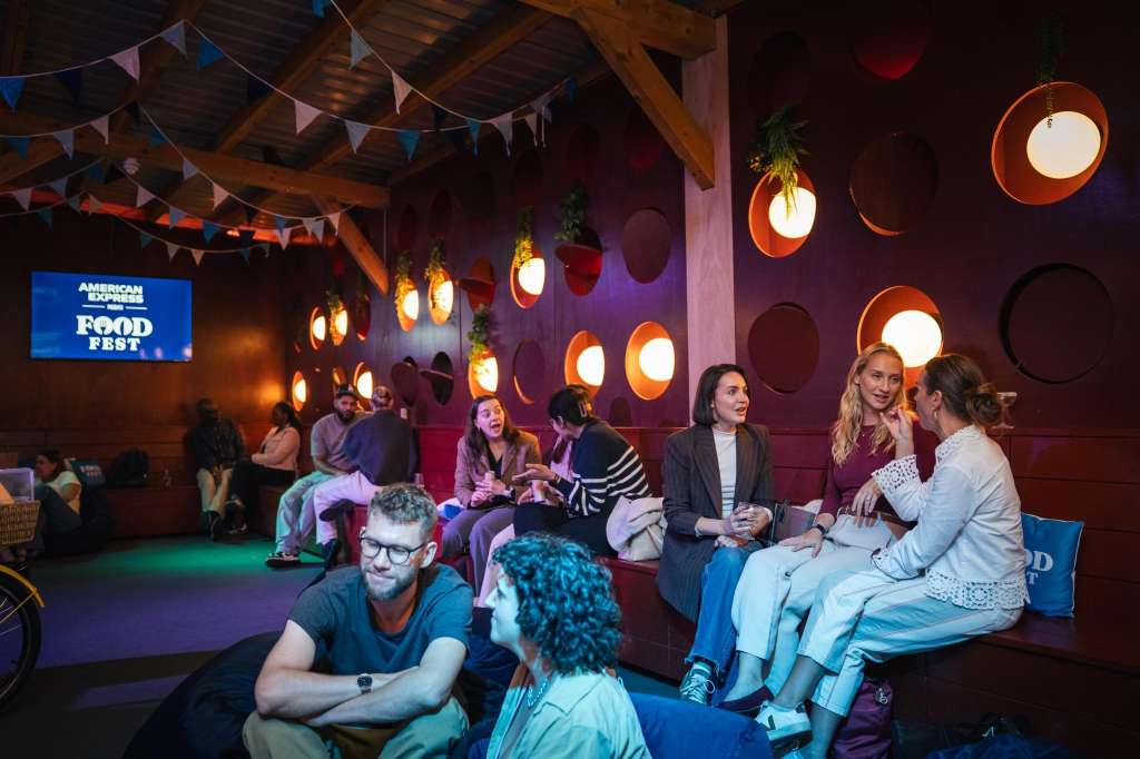 Large industrial venue with exposed wooden beams, colorful bunting, orange pendant lights, and guests mingling during an event.