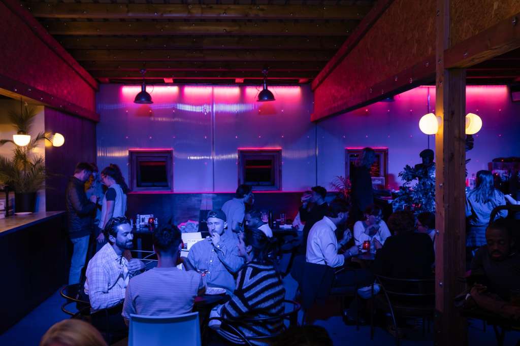 Bar interior with neon pink and blue lighting, multiple guests mingling and seated at tables.