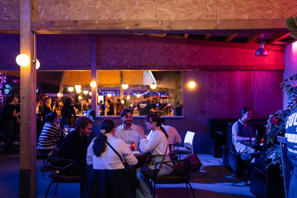 Industrial venue with exposed beams, colorful mood lighting in purple and red, guests mingling at tables and bar area.