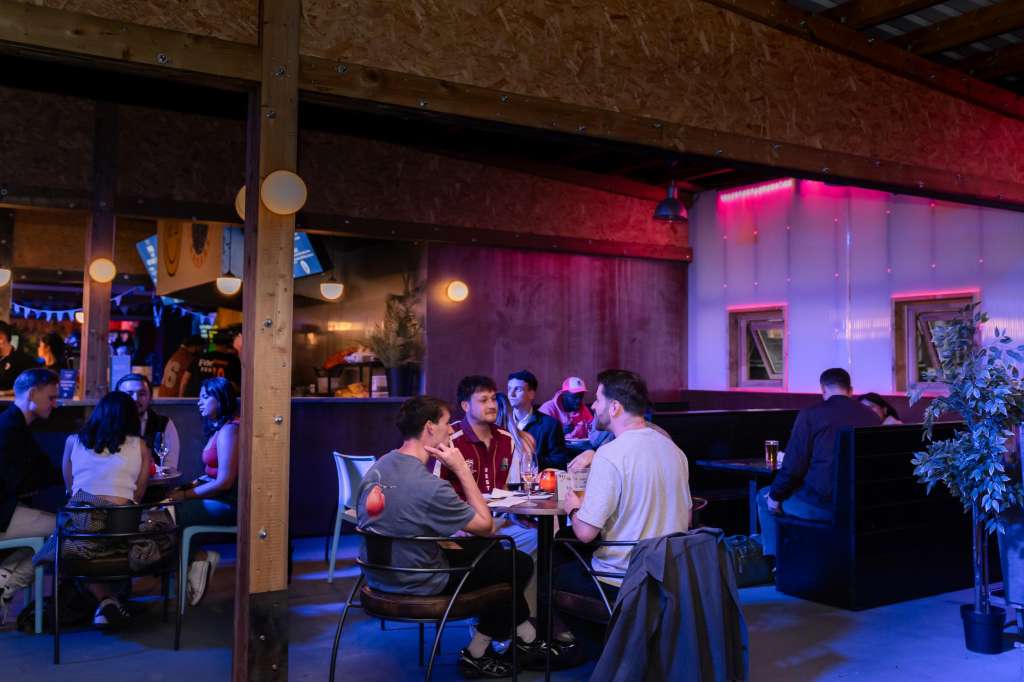 Hotel Milano Bar with guests seated at tables, neon pink and blue lighting, industrial wooden beams, bar counter visible.