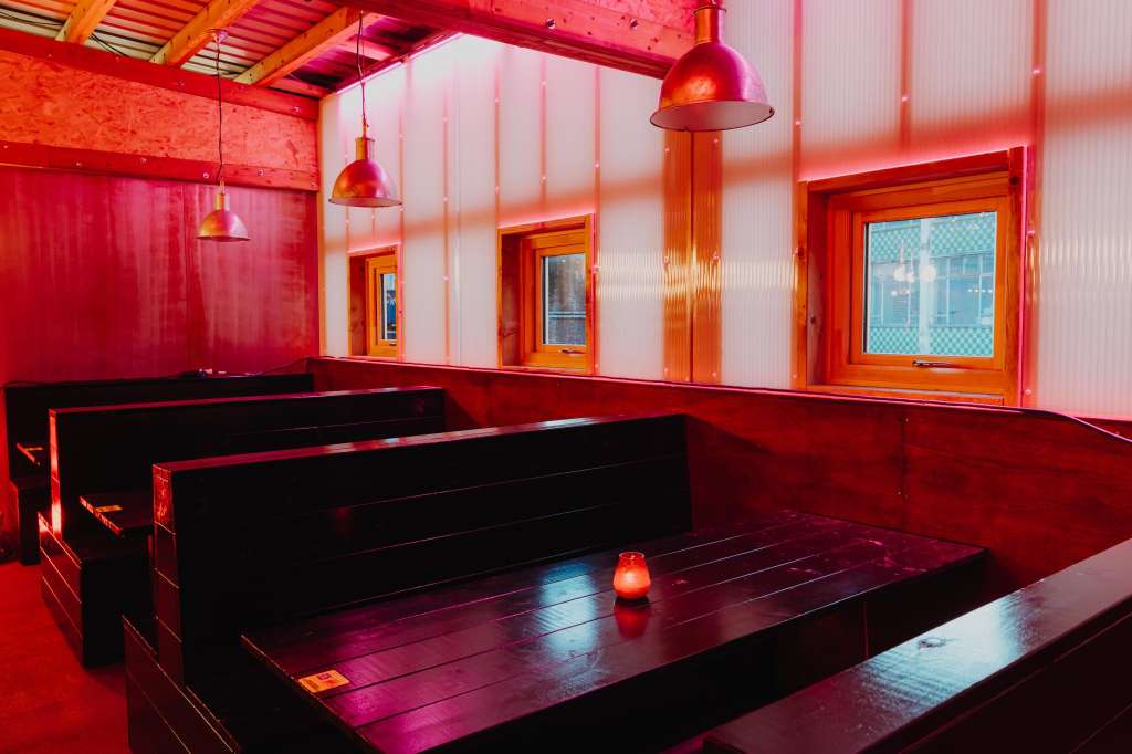 Modern event space with red and orange ambient lighting, black seating platforms, pendant lights, and corrugated metal walls.