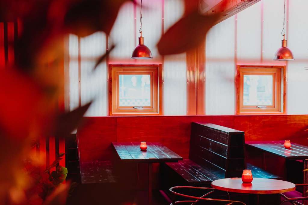 Modern bar interior with red ambient lighting, pendant lamps, round tables with black chairs, and large windows.