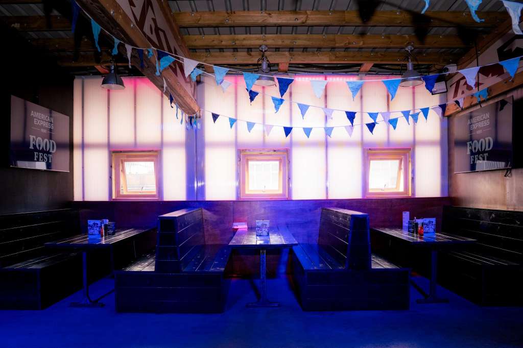 Industrial venue with exposed wooden beams, illuminated white panels, blue bunting, and booth-style seating arranged for an event.