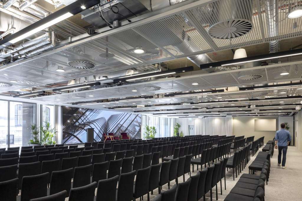 Auditorium with rows of black chairs facing a stage, industrial ceiling, and large windows with city views.