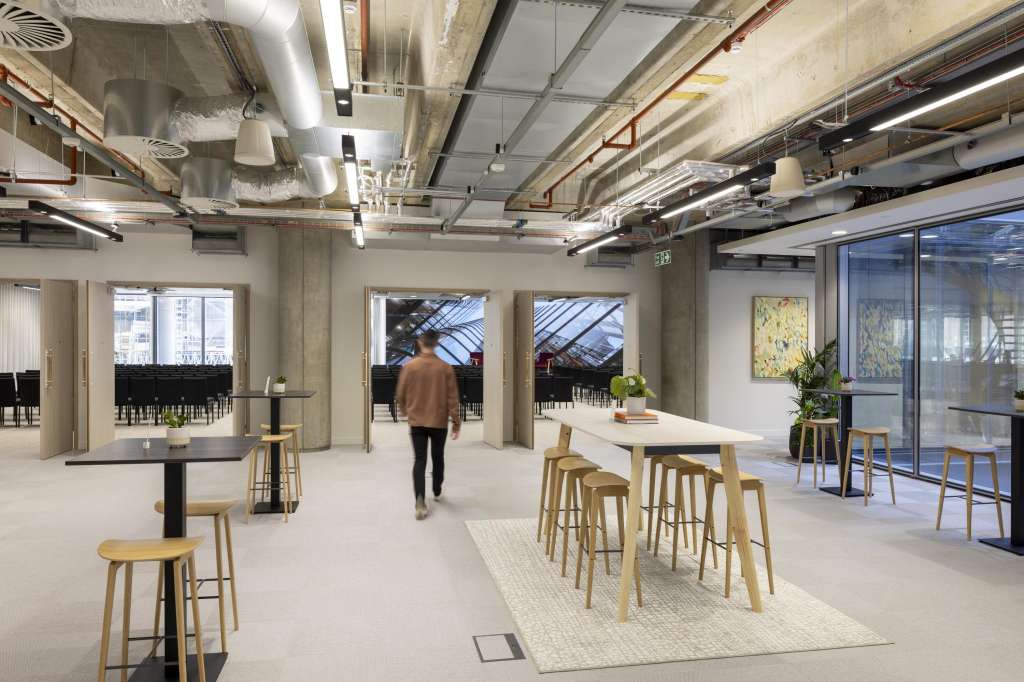 Gallery space with high ceilings, exposed ductwork, bar seating, and large windows overlooking cityscape.