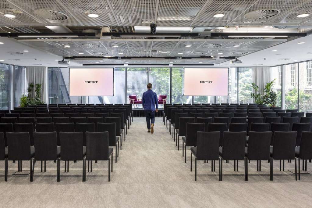 Auditorium with rows of black chairs facing two projection screens displaying TOGETHER branding, one person visible.