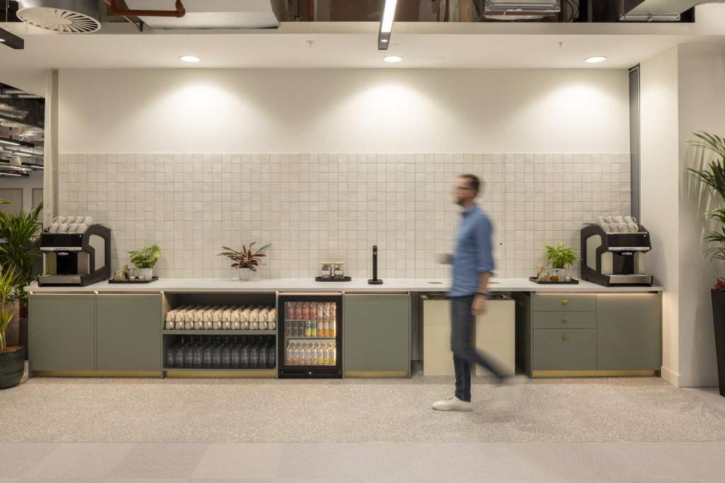 Modern break room with white tile backsplash, sage cabinetry, coffee machines, beverage cooler, and potted plants.