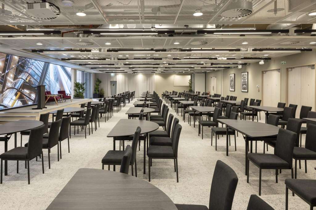 Large open space with multiple round tables, black chairs, and recessed ceiling lighting. Video screen visible on left wall.