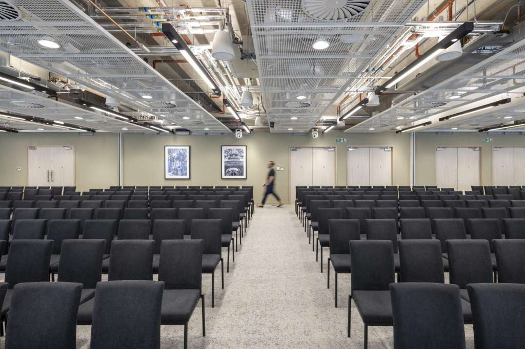 Auditorium with rows of black chairs, industrial ceiling, and framed artwork on walls.