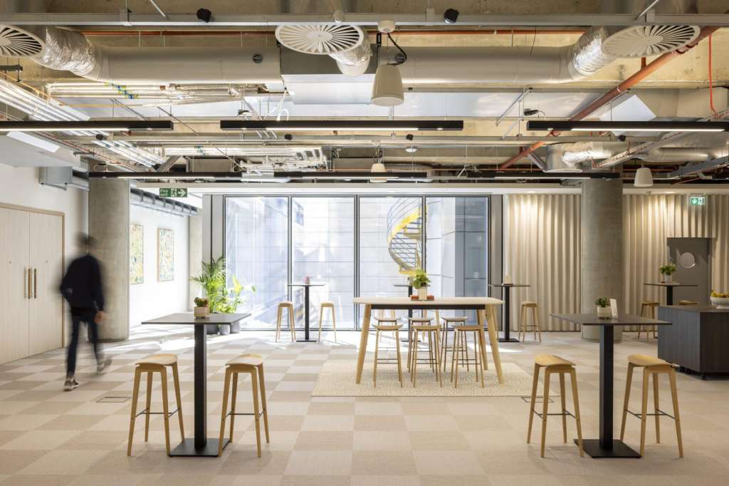 Gallery space with high ceilings, exposed ductwork, wooden bar tables, stools, and large windows overlooking the street.