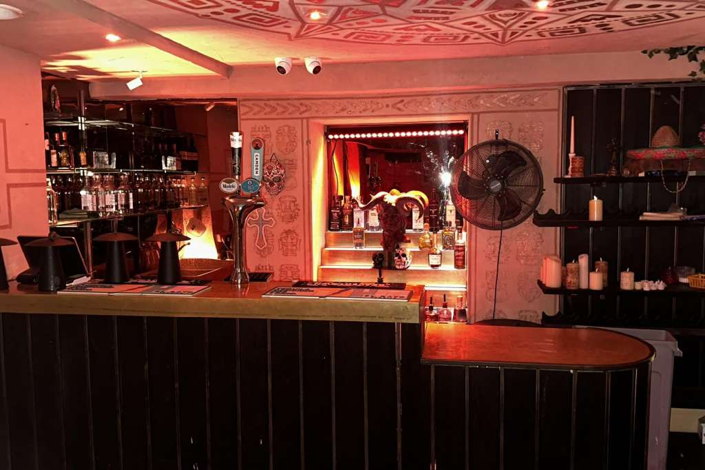 Bar counter with black paneling, warm ambient lighting, shelving with bottles and decor, mirror backdrop.