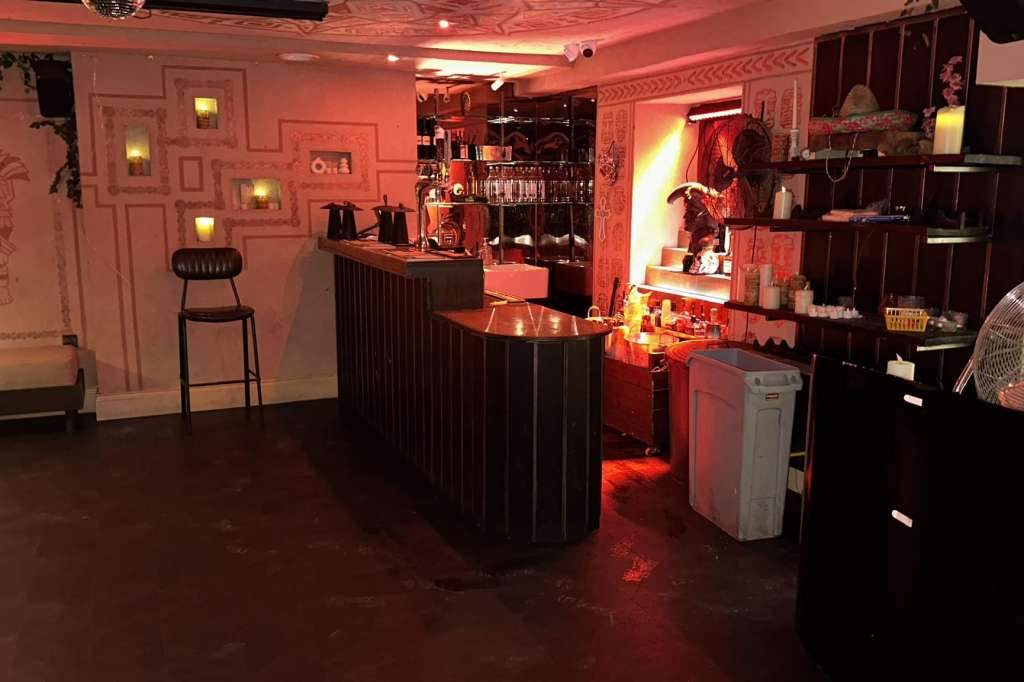Bar counter with red ambient lighting, shelving, and decorative wall panels in an empty venue space.