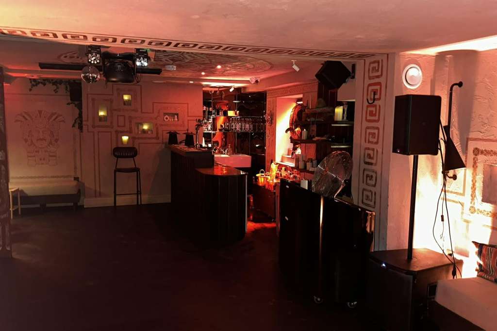 Modern event space with red ambient lighting, bar counter, high-top tables, and professional sound equipment.