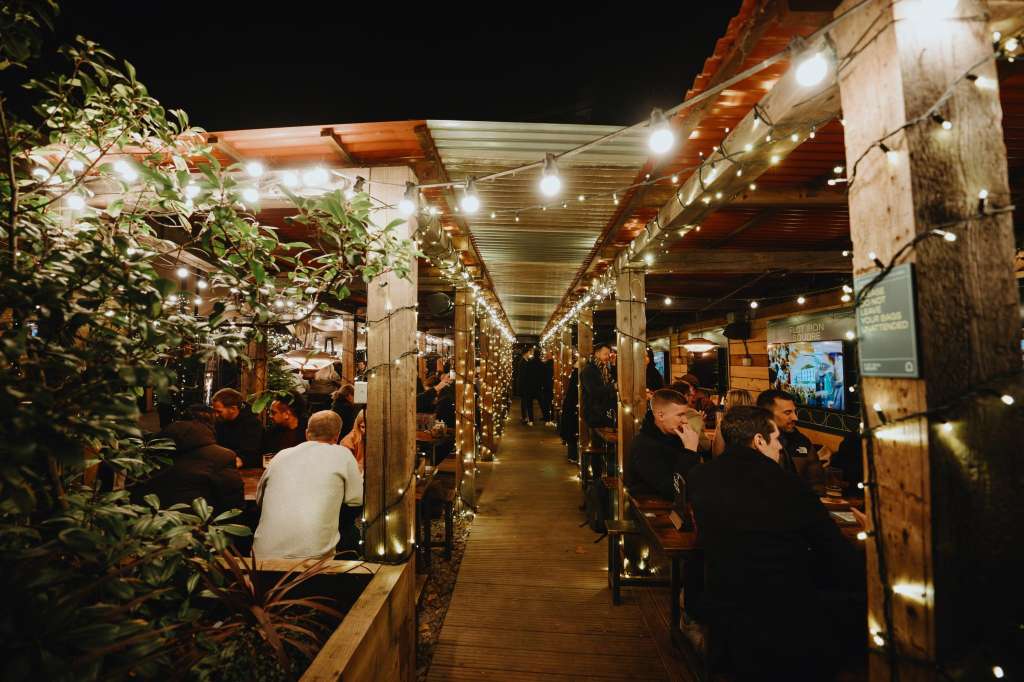 Terrace with string lights, wooden pergola, potted plants, and guests mingling in evening setting.