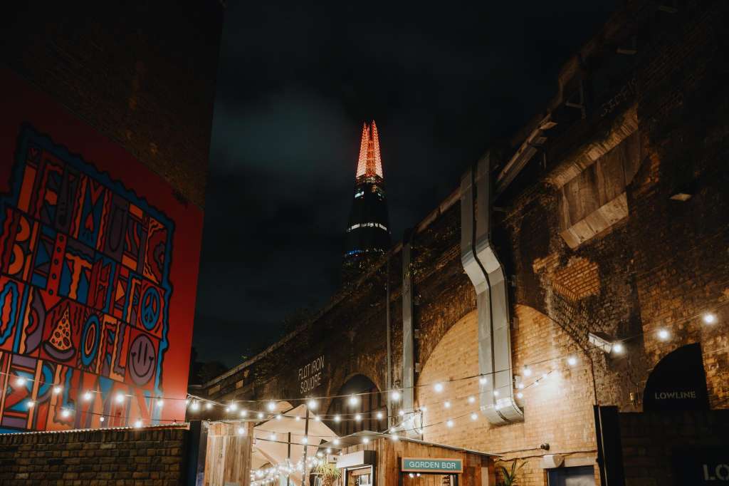 Outdoor terrace at night with string lights, brick walls, and illuminated signage overlooking a city skyline.