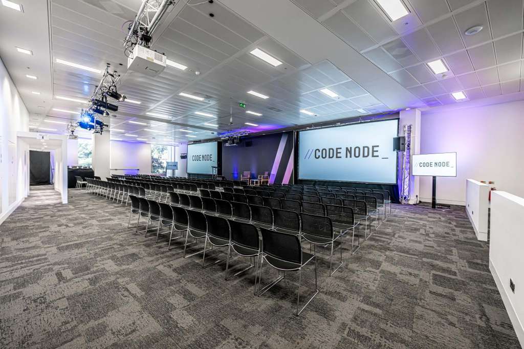 Conference room with theater-style seating, projection screens, stage lighting, and CODE NODE branding on walls.