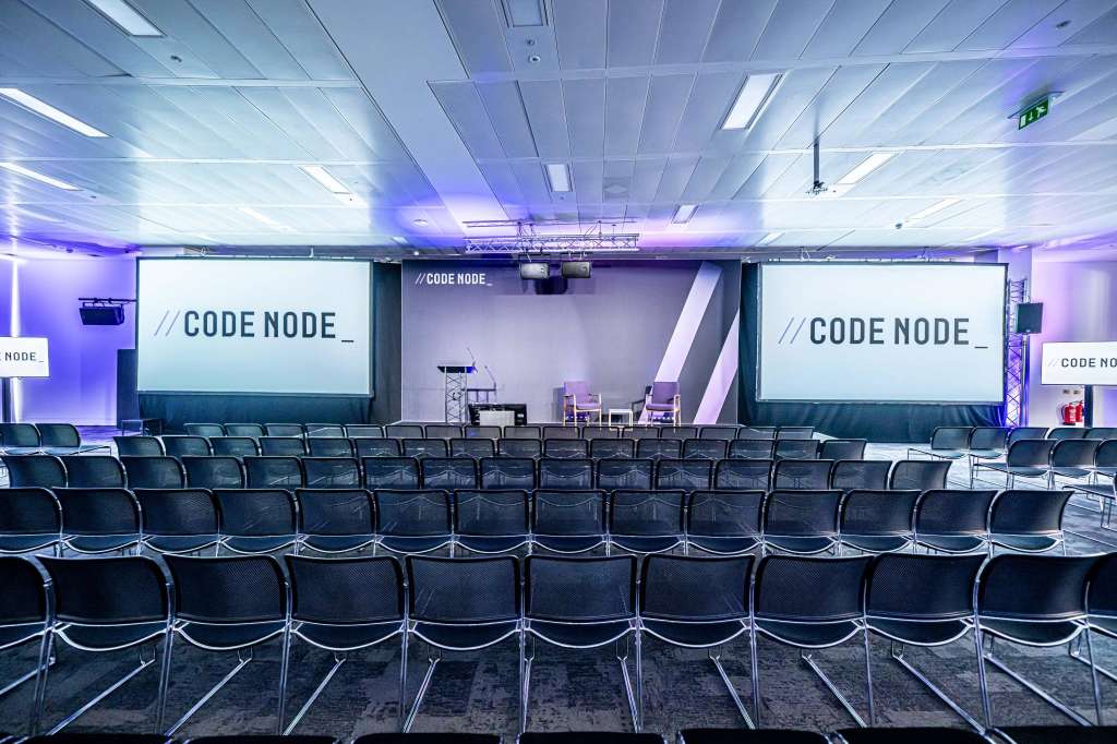 Large auditorium with rows of black chairs facing two projection screens and a stage, purple ambient lighting throughout.