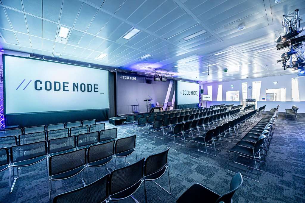Large auditorium with rows of black chairs facing projection screens and stage with blue ambient lighting.