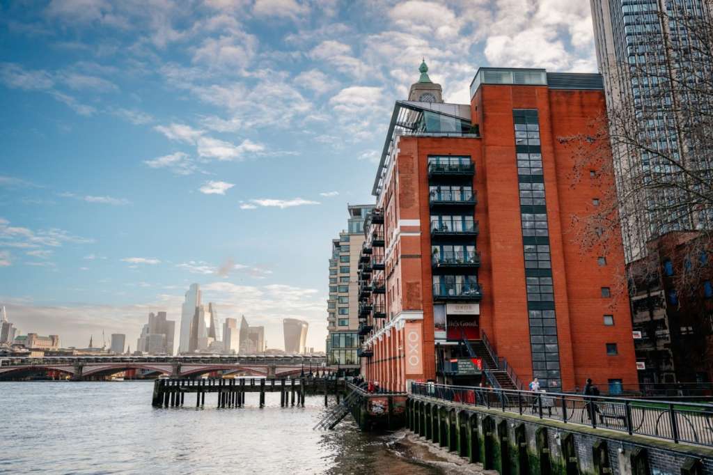 Exterior view of red-brick building on Thames riverfront with London skyline and bridge in background.