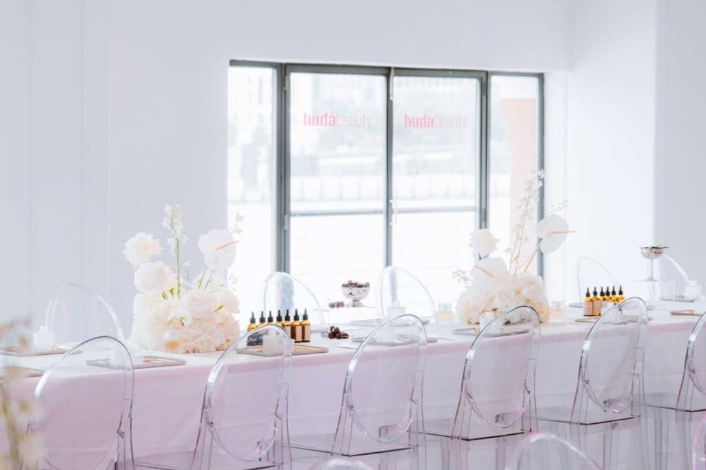 Long table with pink linens, clear chairs, white floral arrangements, and product displays by large windows.