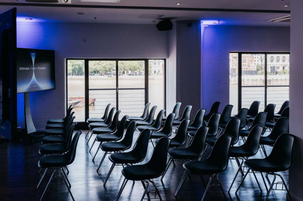 Modern conference room with rows of black chairs facing large windows overlooking a plaza, blue accent lighting on walls.