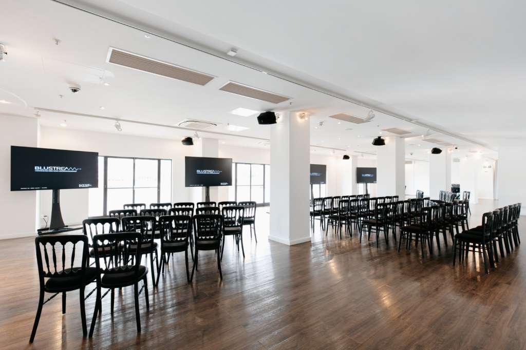 Modern event space with black chairs arranged in rows, multiple screens displaying branding, and wooden flooring.