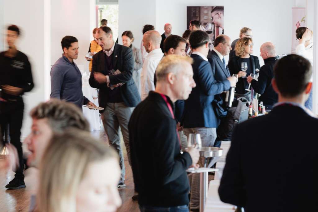 Modern gallery space with standing guests mingling, holding drinks, white walls, track lighting, and artwork on display.