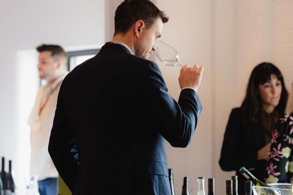 Man in blue suit drinking from glass at table with wine bottles and guests in background.