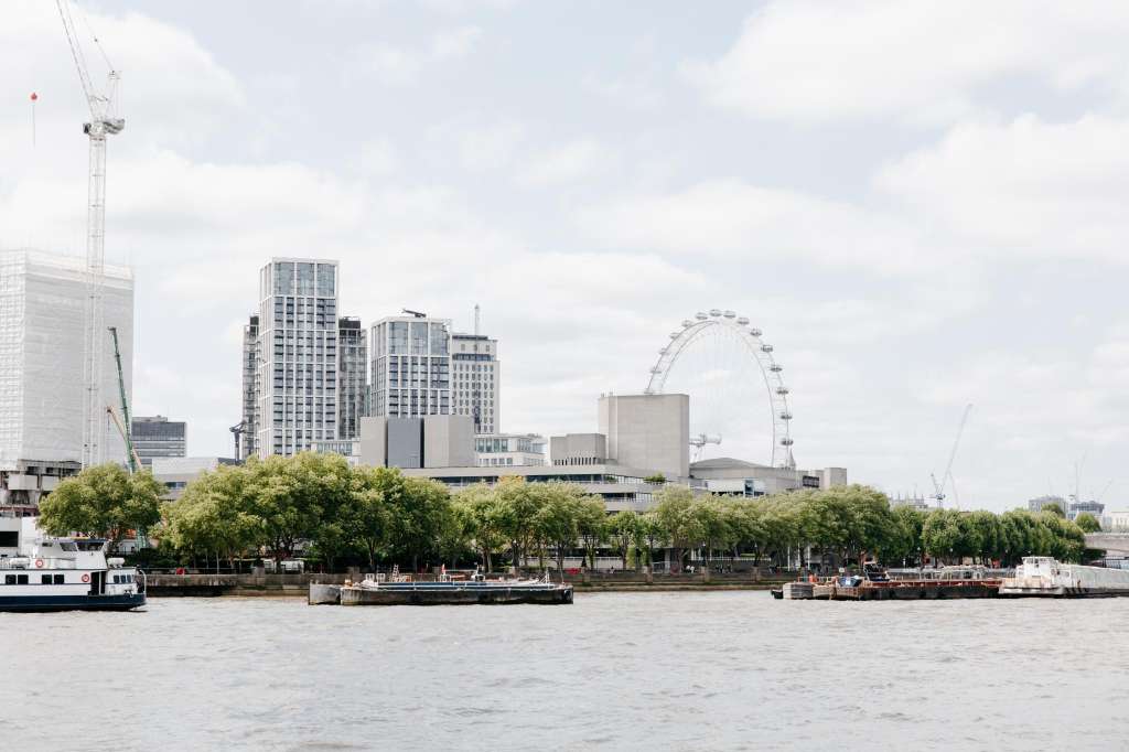 Exterior view of riverside cityscape with modern buildings, trees, and observation wheel across water.