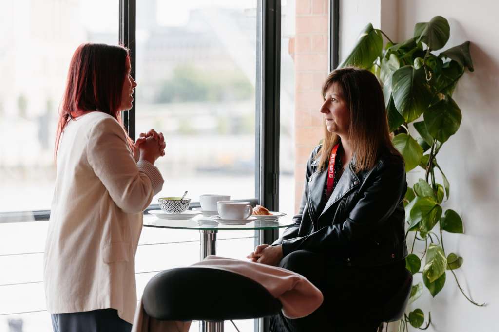 Two people having a conversation at a small table by floor-to-ceiling windows with city views and potted plant.