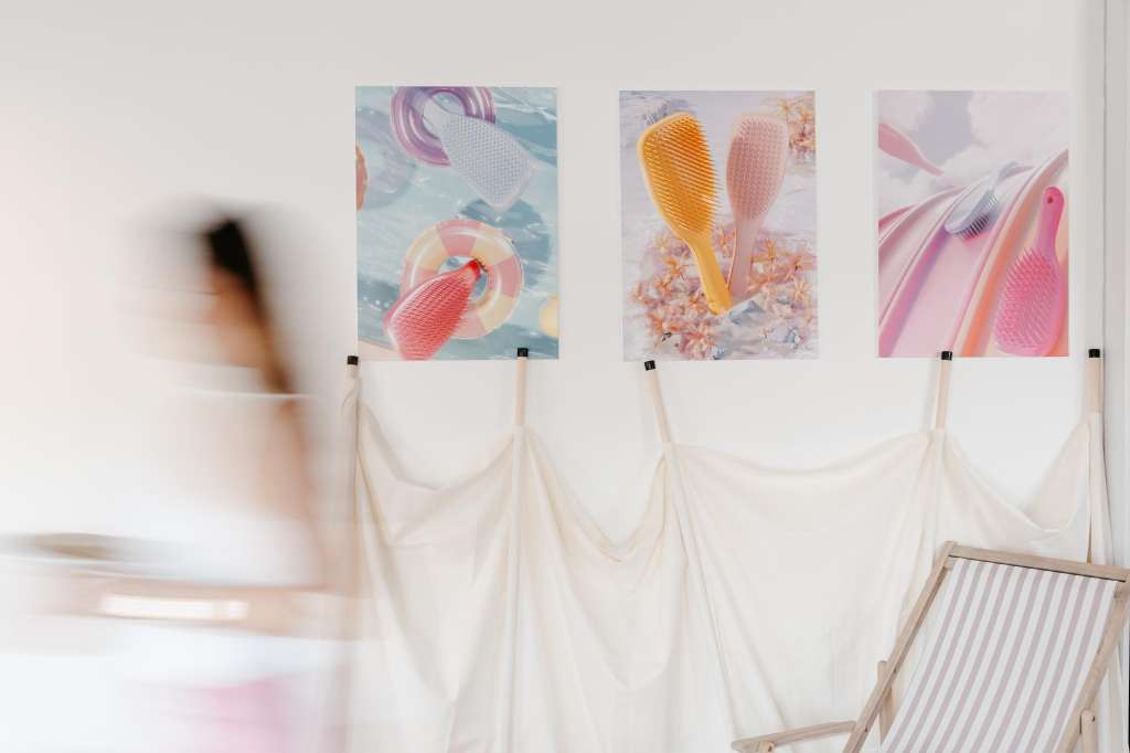 Exhibition wall with four framed prints of colorful beauty products displayed above cream fabric draping and a striped chair.