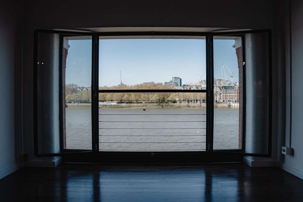 Empty modern room with floor-to-ceiling windows overlooking a river, cityscape, and moored boats.