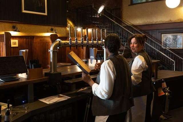 Bar interior with brick walls, globe pendant lights, beer taps, and staff in waistcoats behind counter.