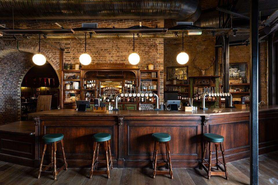 Bar counter with wooden paneling, pendant lighting, shelved spirits display, and teal bar stools in industrial brick interior.