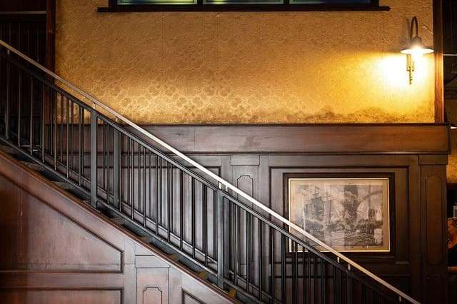 Staircase with wood paneling, decorative windows, round tables with green stools, and warm lighting.