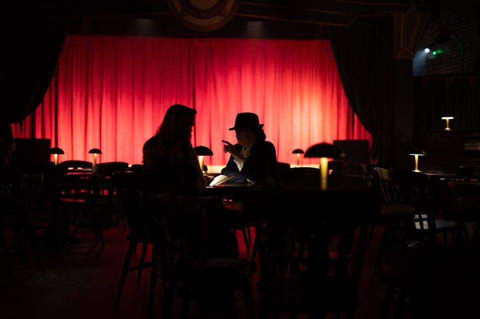Two people seated at a table against a dramatic red curtain backdrop with ambient lighting and small table lamps.