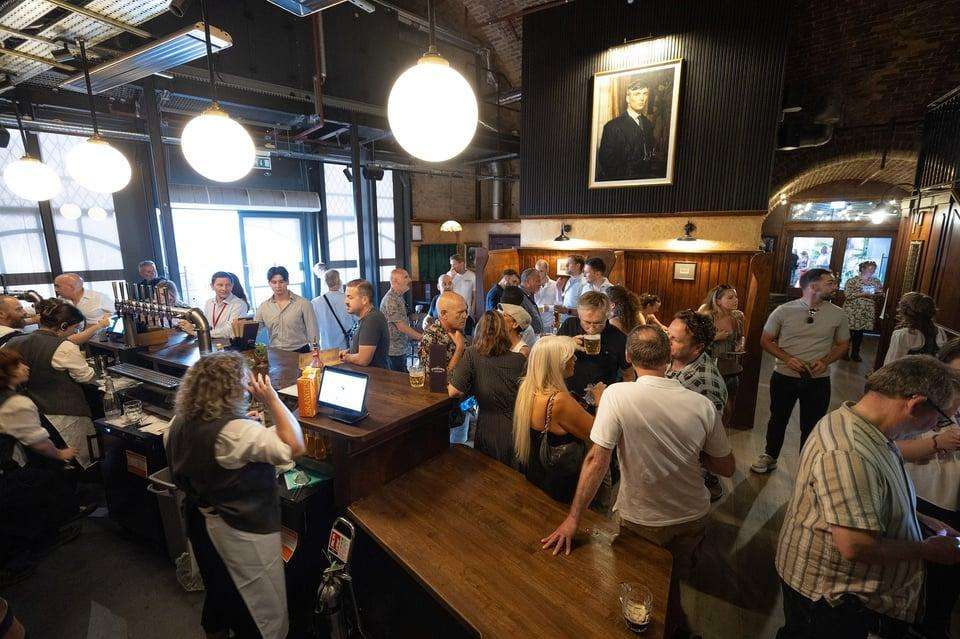 Industrial tavern interior with standing guests, wooden bar, pendant lighting, and exposed brick walls.