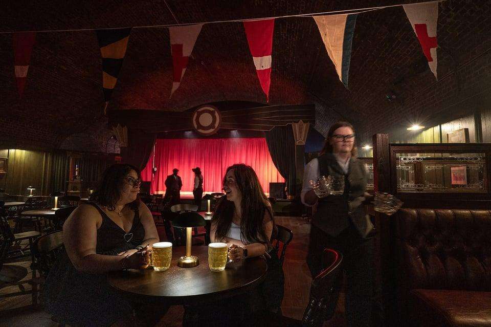 Interior bar space with red stage lighting, bunting, round tables with drinks, and guests mingling.