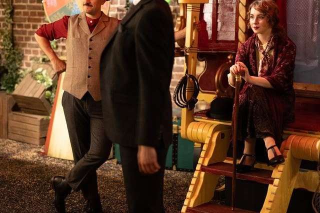 Three people in period costume interact near vintage fairground equipment and exposed brick wall with decorative bunting.